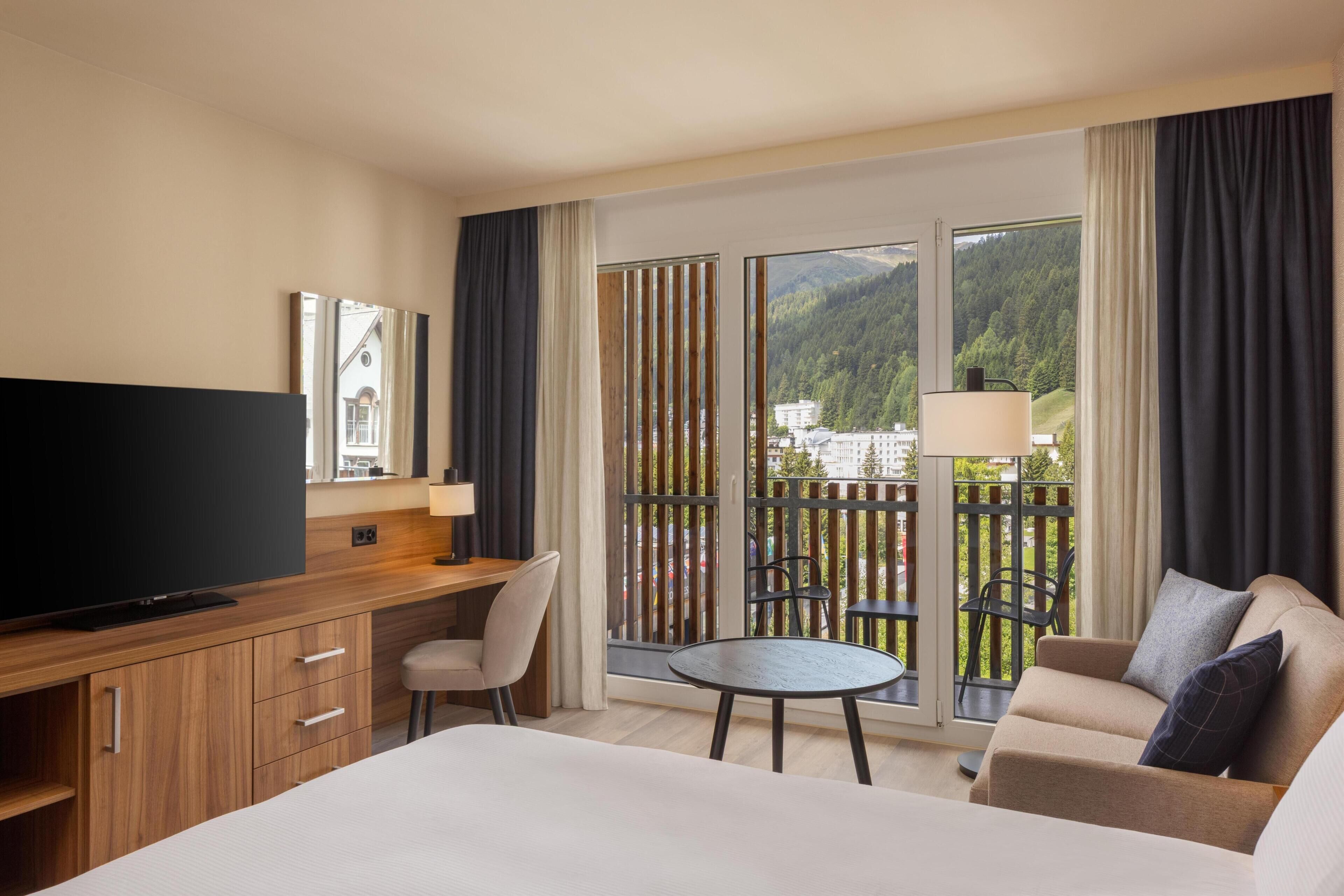 Habitación de hotel con cama, escritorio con televisor, una silla, una mesita y balcón con vista a las montañas.