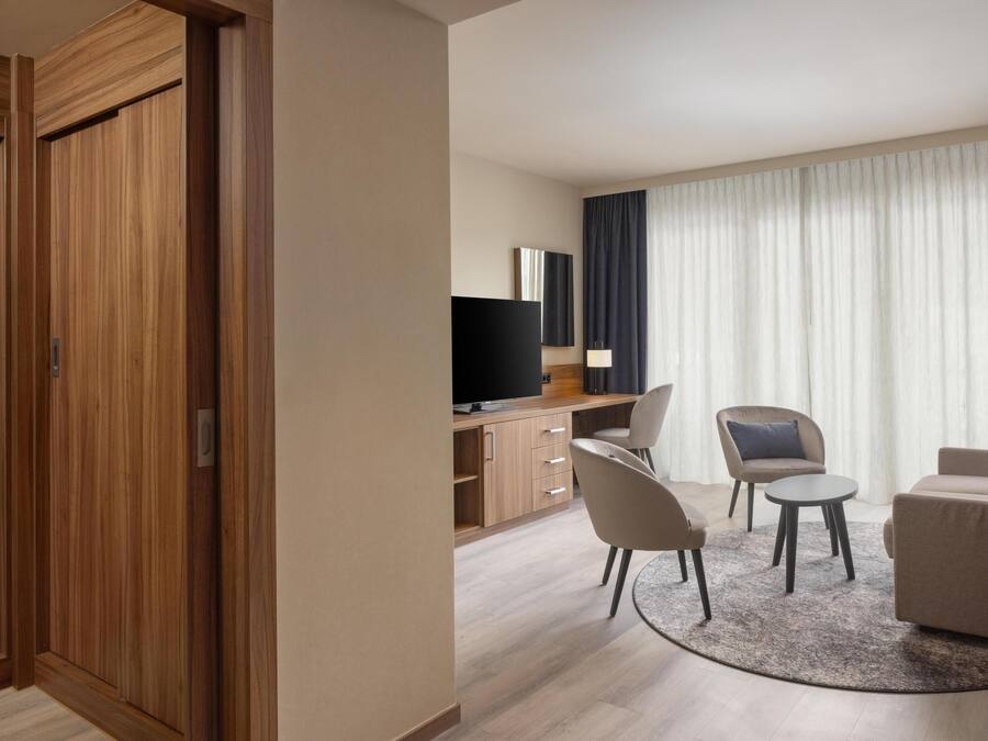 Habitación de hotel con armario de madera, televisor, escritorio, silla, sofá y un amplio ventanal con cortinas.