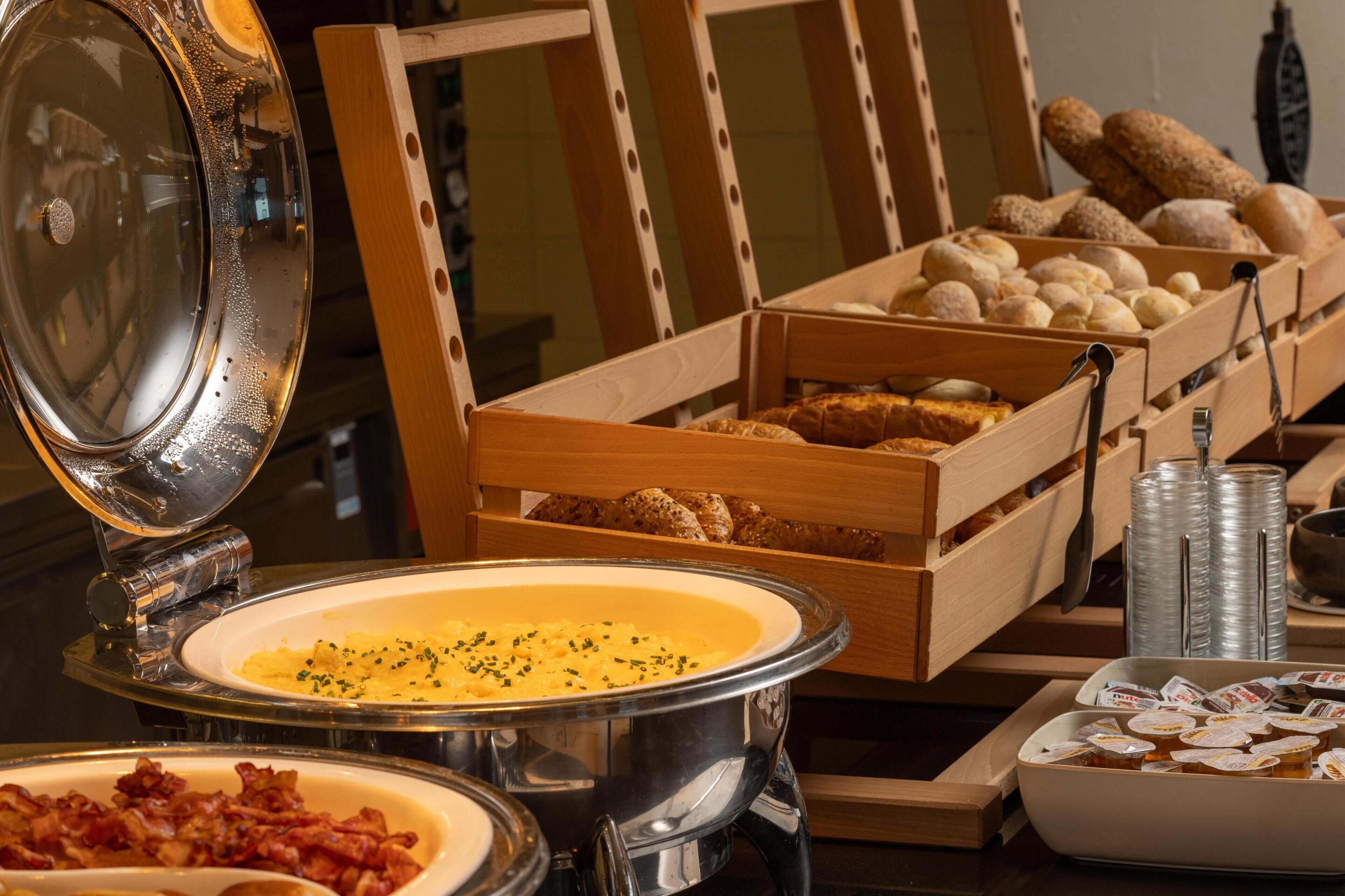 A buffet setup with various food items, including a bowl of soup, a container of bacon, and multiple boxes of bread.
