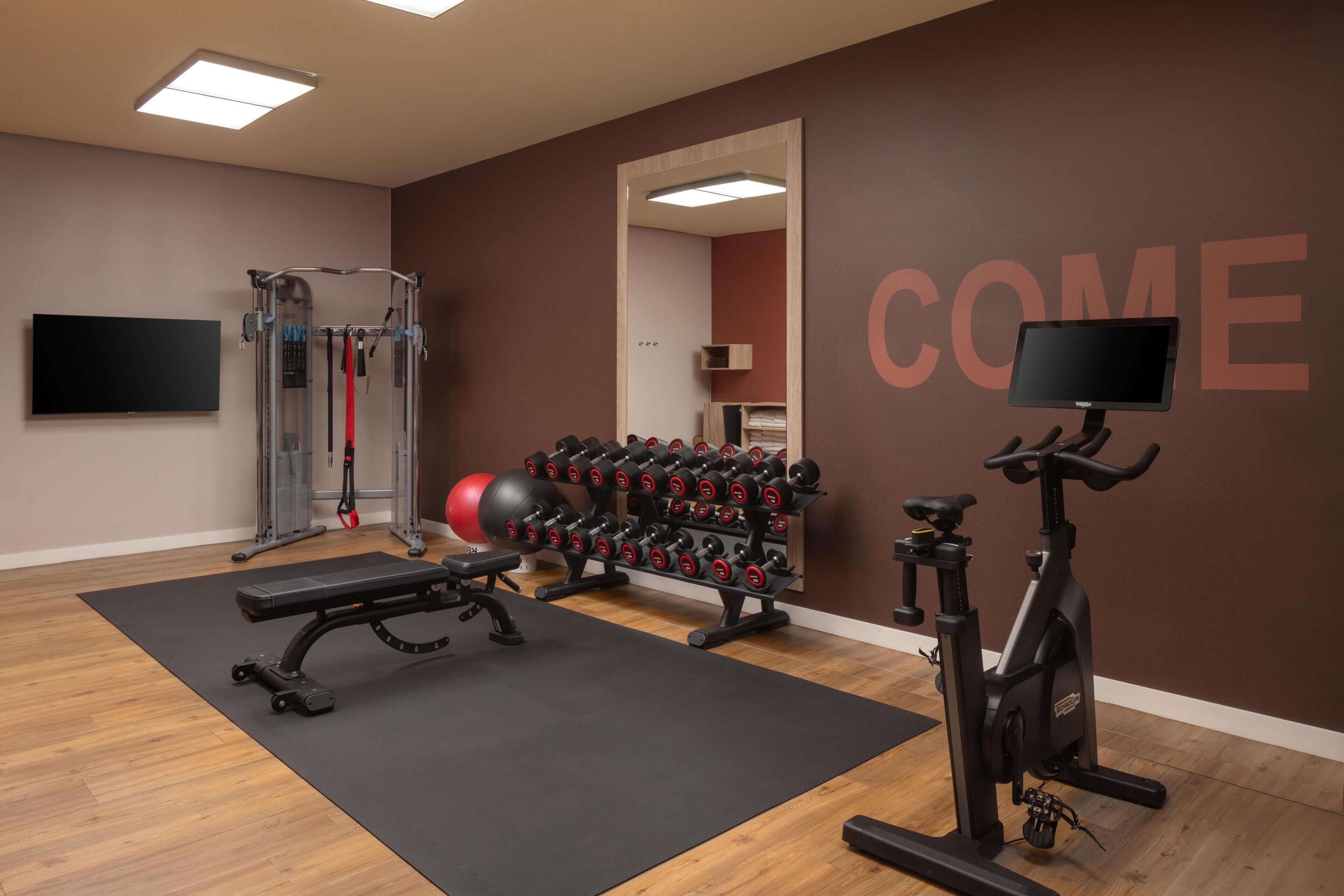 A gym with exercise equipment, a wall-mounted TV, and a motivational 'COME' sign.