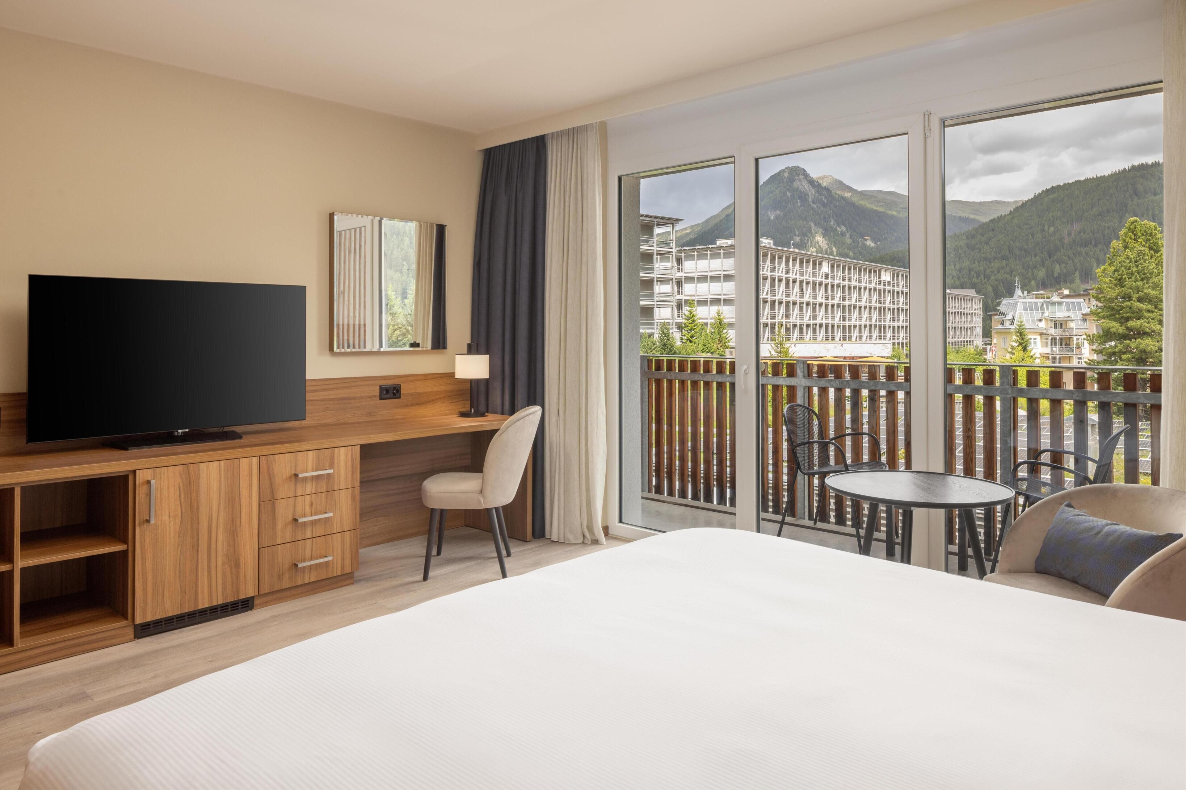 Habitación de hotel con una cama grande, televisión, escritorio con silla y balcón con mesa y sillas.