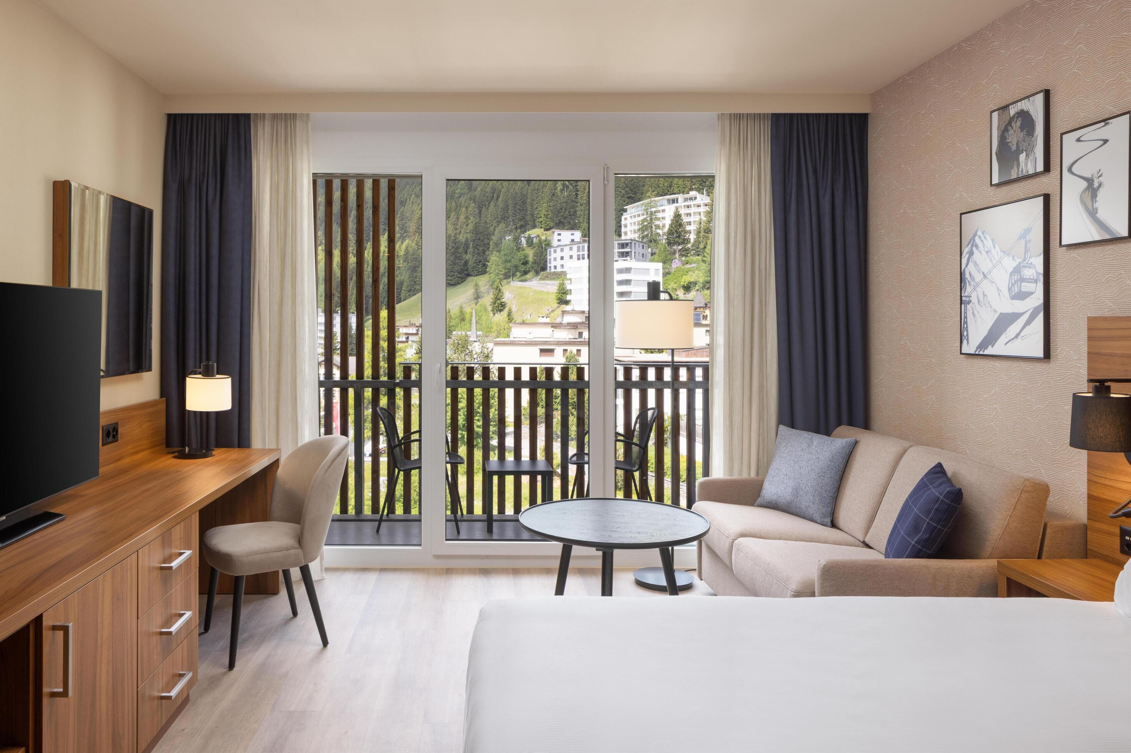 A hotel room with a bed, a sofa, a desk with a TV, a balcony with chairs, and a view of buildings.