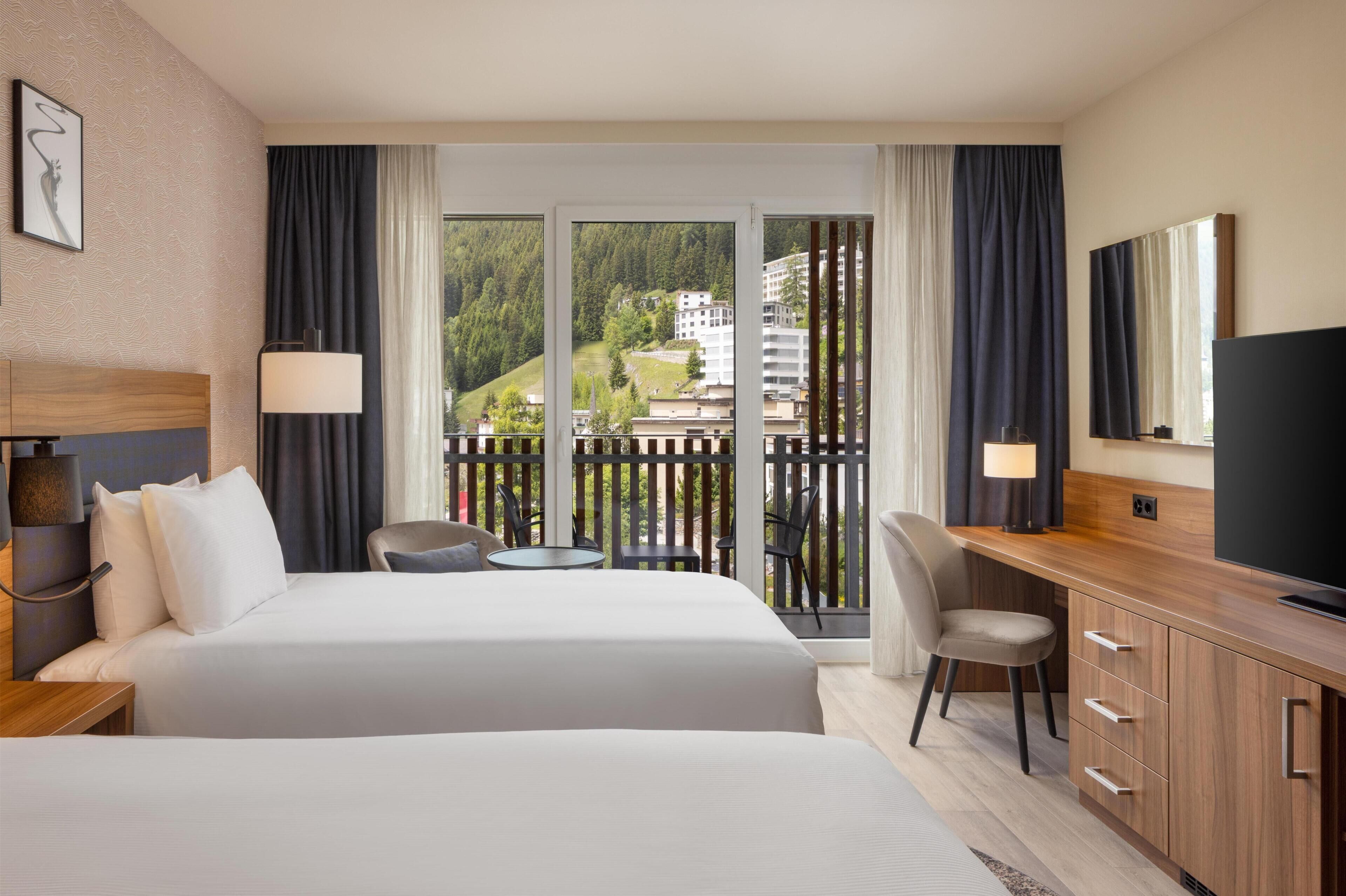 A hotel room with two beds, a desk, a TV, and a balcony with a view of buildings and greenery.