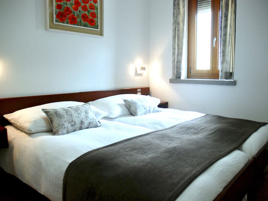 A neatly made bed with a grey blanket and two pillows, a wooden headboard, and a painting of red flowers on the wall.
