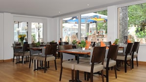 A restaurant with wooden tables and chairs, large windows with views of the outside, and indoor plants.