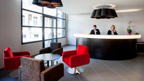 A modern reception area with a curved desk, red and brown chairs, and large windows providing natural light. - Hotel Panorama (Lourdes)