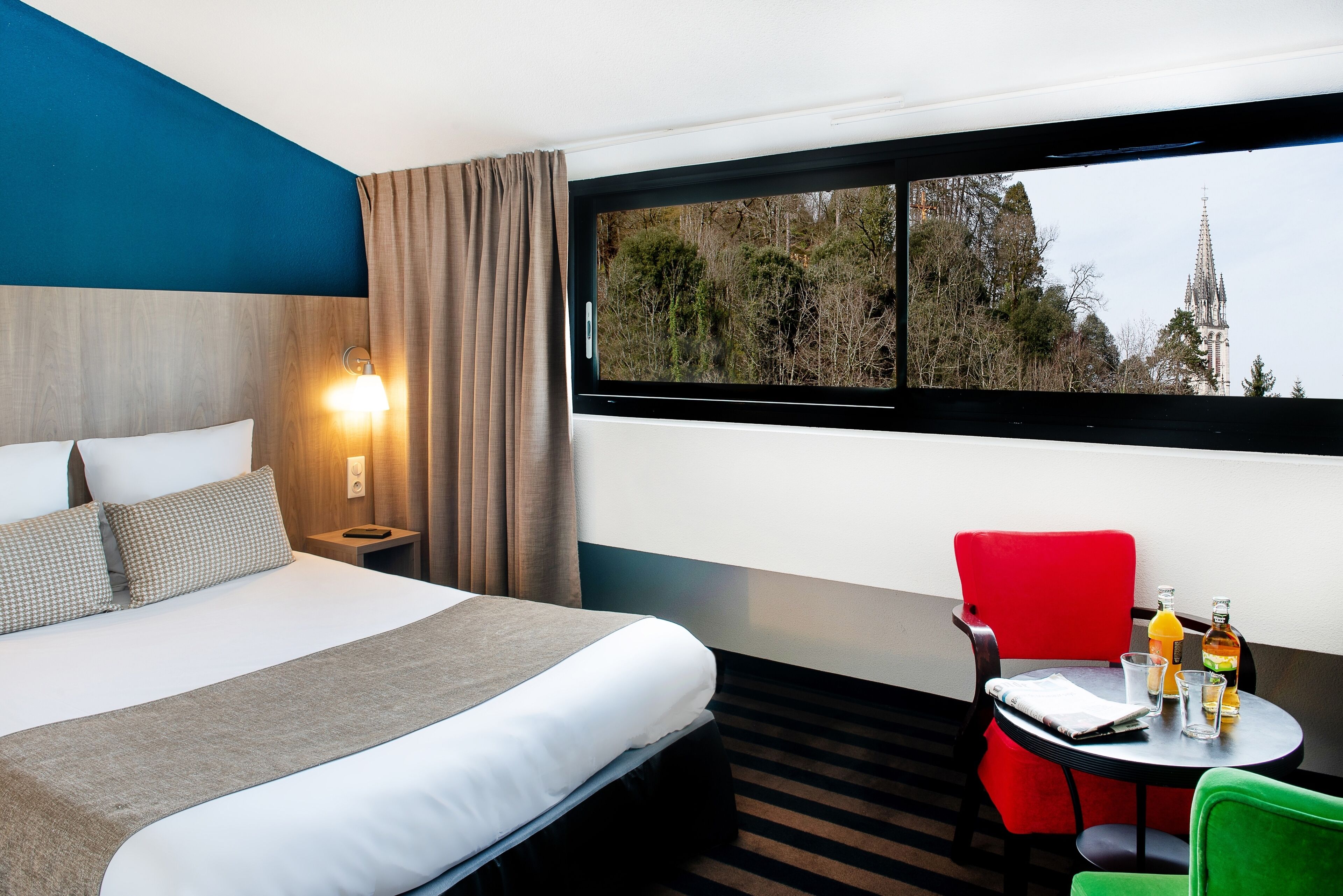 A hotel room with a bed, a table with drinks, a red chair, and a window with a view of trees and a building.