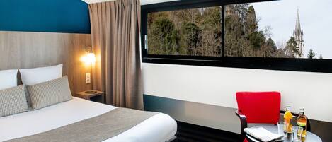 A hotel room with a bed, a table with drinks, a red chair, and a window with a view of trees and a building.