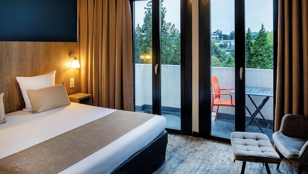 A hotel room with a bed, a nightstand, a chair, and a view of the outdoors through a sliding glass door. - Hotel Panorama (Lourdes)