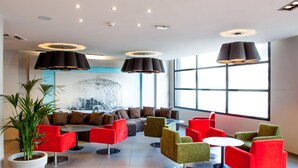 A modern lounge area with multiple seating options, including red and green chairs, a sofa, and a round table. There are large windows providing natural light and a potted plant adding greenery. - Hotel Panorama (Lourdes)