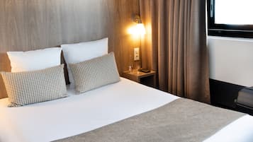 Una cama bien hecha con dos almohadas, una mesita de noche con una lámpara y un ventanal con cortinas.