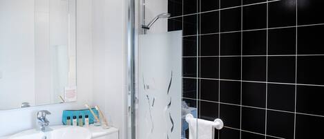 Un baño con lavamanos blanco, cabina de ducha con mampara de vidrio y paredes cubiertas de azulejos negros.
