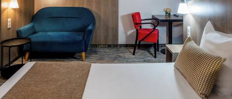A hotel room with a bed, a blue armchair, a red chair, a small table, and two wall-mounted lamps.