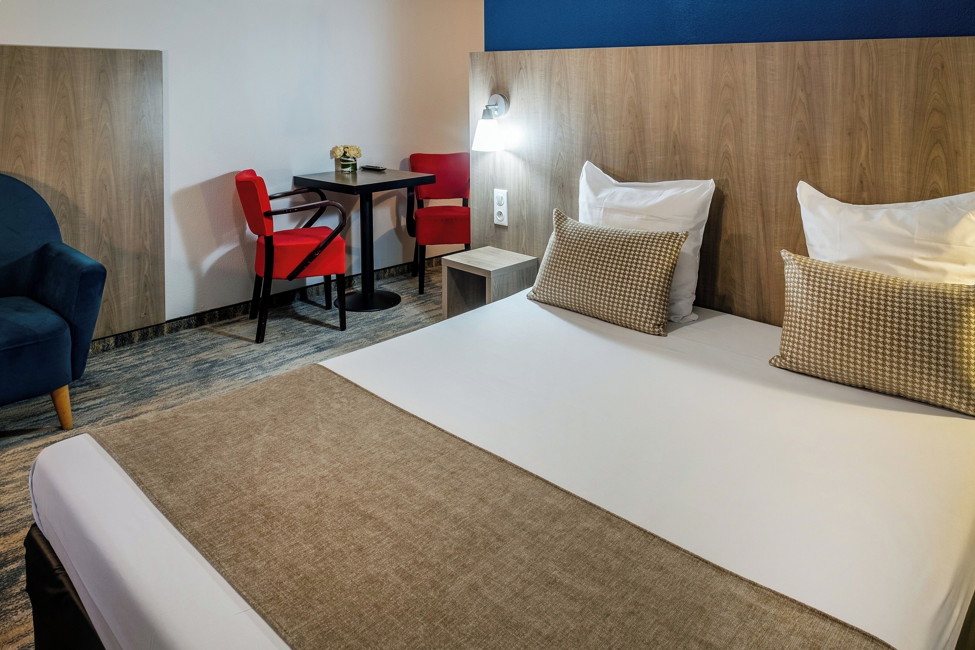 Una habitación de hotel con una cama, dos sillas rojas, una mesita y una lámpara.