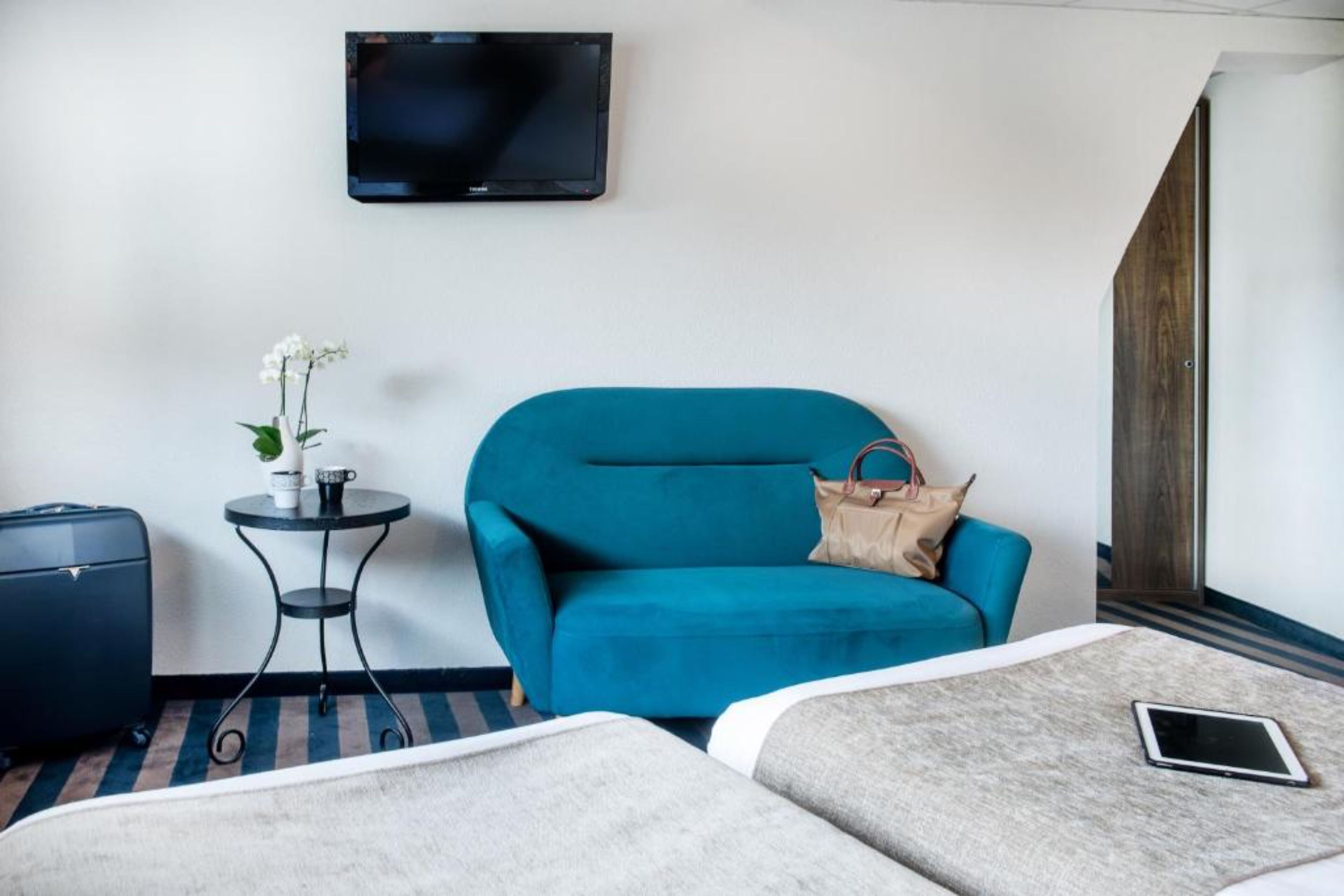 A hotel room with a teal sofa, a small round table with a plant, a TV mounted on the wall, a suitcase, and two beds.