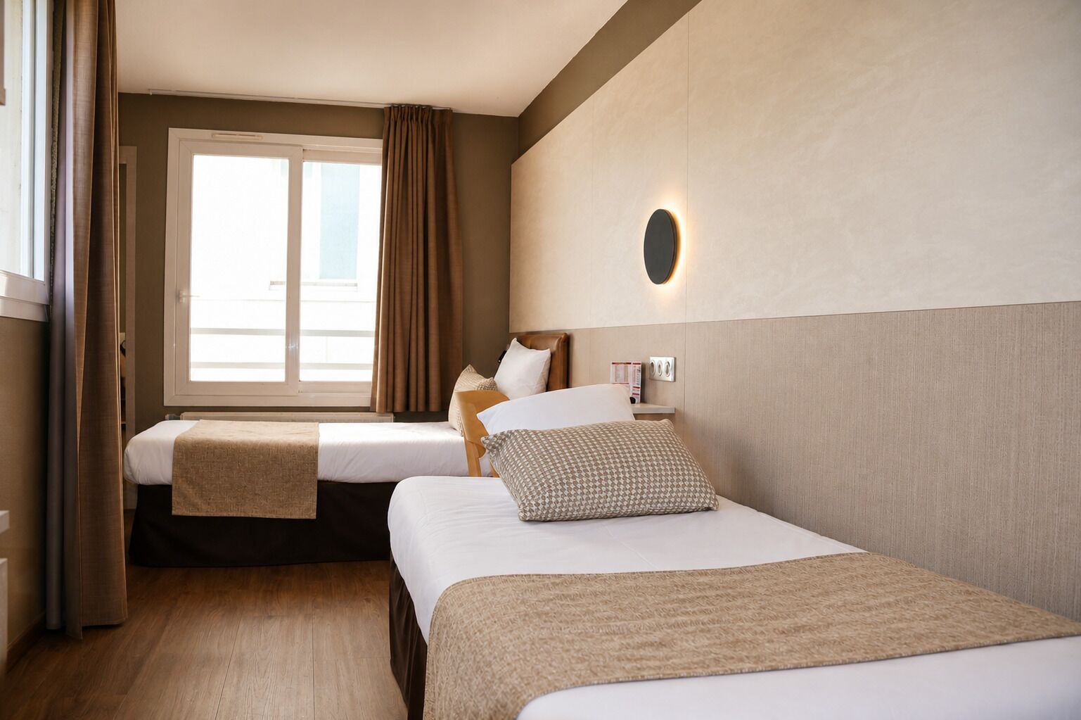 A hotel room with two beds, a window with curtains, a wall-mounted light, and a wooden floor.
