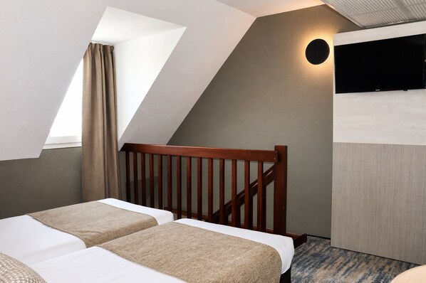 Une chambre avec un plafond en pente, une tête de lit en bois, deux lits avec une literie beige, un petit téléviseur fixé au mur et un luminaire circulaire.