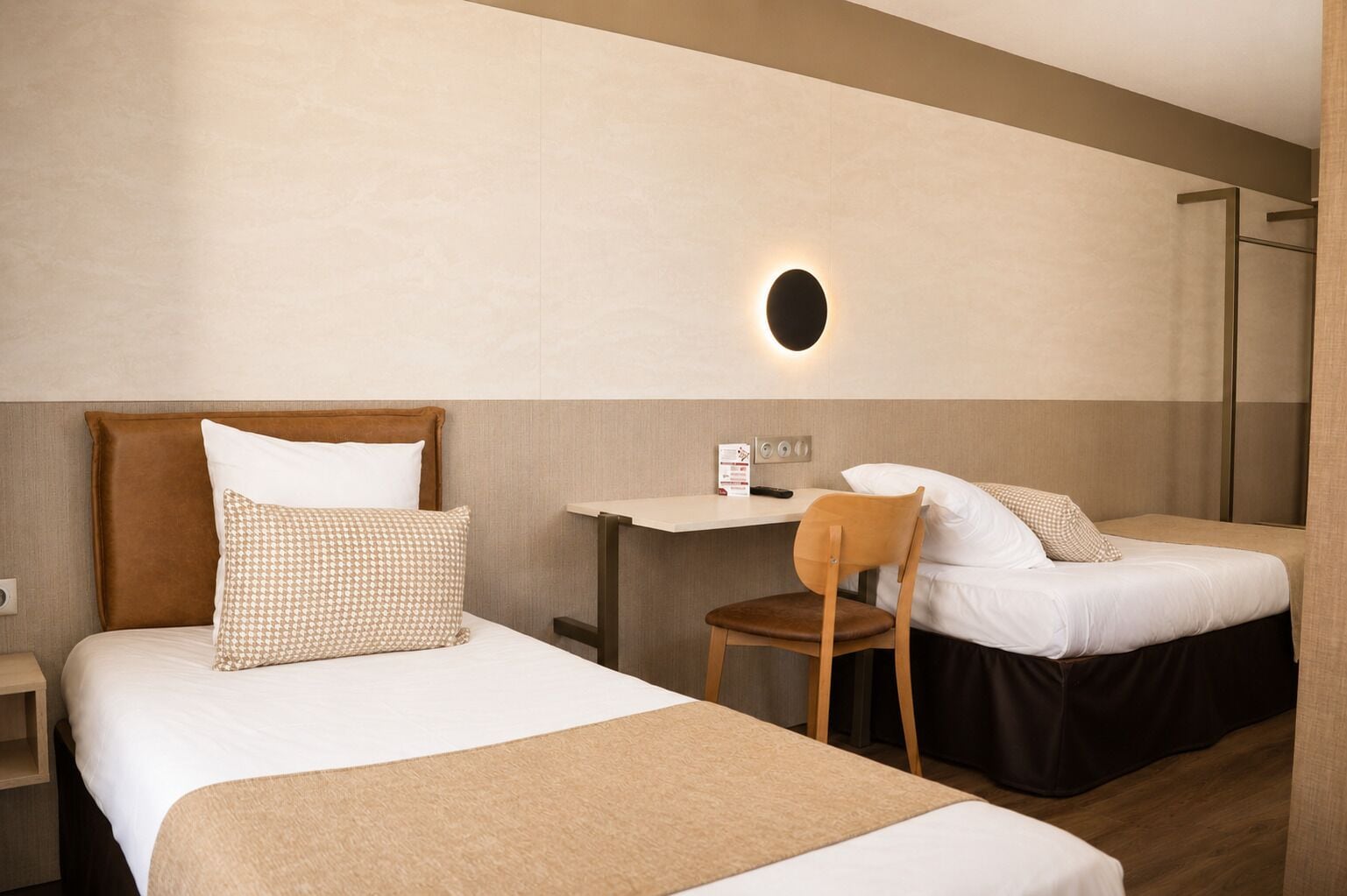 A hotel room with two beds, a desk, a chair, and a wall-mounted light.