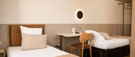 A hotel room with two beds, a desk, a chair, and a wall-mounted light.