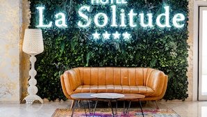 A room with a leather sofa, a round coffee table, a decorative lamp, and a wall with a neon sign reading "HOTEL La Solitude". - Hotel La Solitude (Lourdes)