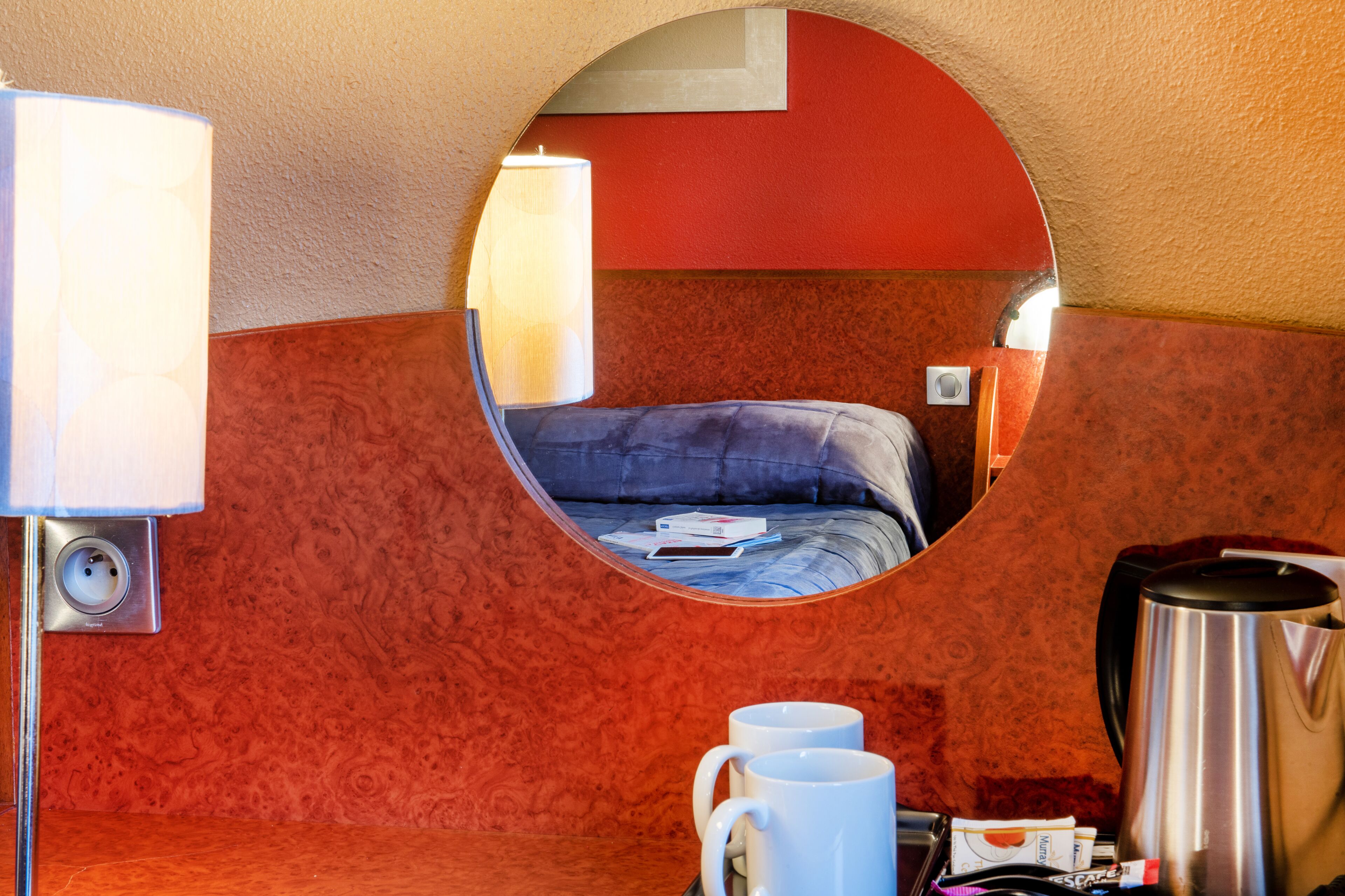 A hotel room with a circular mirror, a bed, a coffee maker, and a nightstand with a lamp and a cup.
