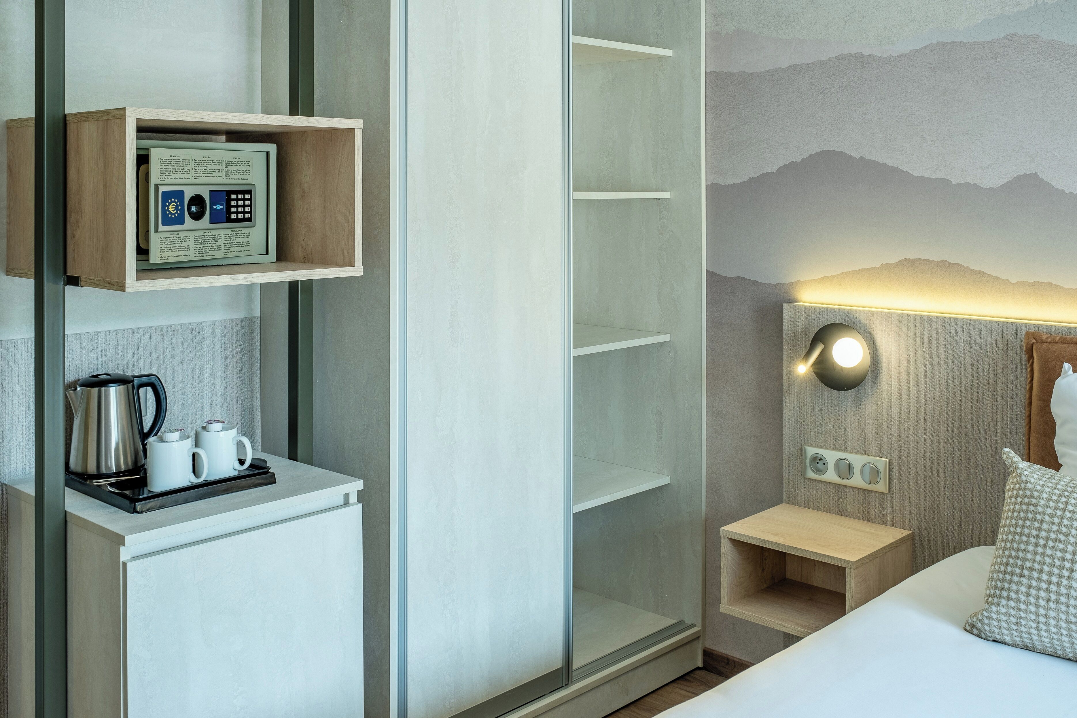 Une chambre d’hôtel comprenant un lit, une table de chevet avec une lampe, une étagère avec une machine à café et une station de recharge fixée au mur.