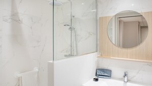 A modern bathroom with a glass-enclosed shower, a white marble wall, a round mirror, and a white sink. - Hotel Saint Sauveur (Lourdes)