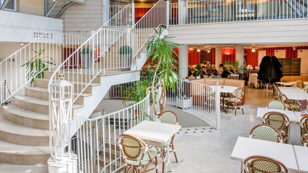 A multi-level indoor space with a central staircase, seating areas, and potted plants. - Hotel Saint Sauveur (Lourdes)