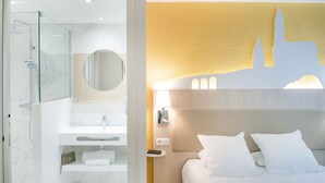 A hotel room with a glass-enclosed shower, a white sink, a mirror, a bed with white bedding, and a wall-mounted lamp. - Hotel Saint Sauveur (Lourdes)