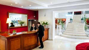 A hotel reception area with a wooden reception desk, a large mirror, and a red wall. There is a staircase with a railing leading to an upper floor. - Hotel Saint Sauveur (Lourdes)