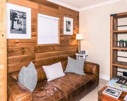 A brown leather sofa with patterned cushions, a wooden wall with framed photographs, a side table with a lamp, and a small shelf with decorative items.