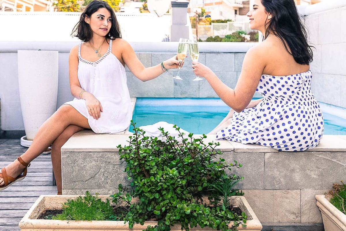 Two women toasting with champagne glasses by a poolside, with a cityscape in the background.