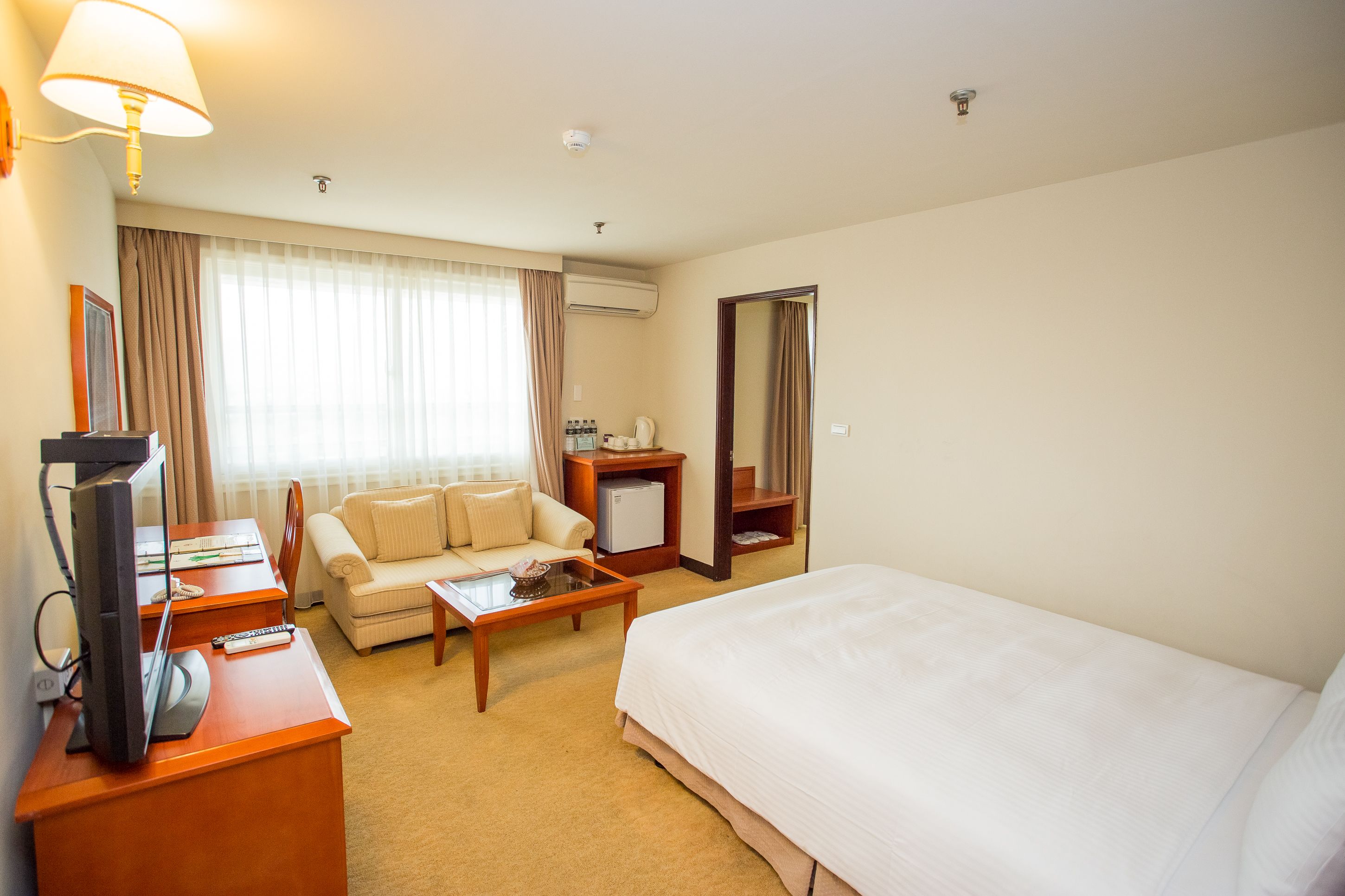 A hotel room with a bed, a sofa, a desk with a computer and a lamp, a coffee table, and a TV.