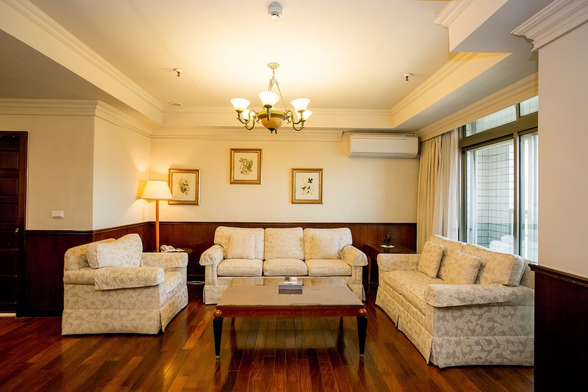 A living room with a sofa set, a coffee table, a chandelier, framed artwork, and a large window with curtains.