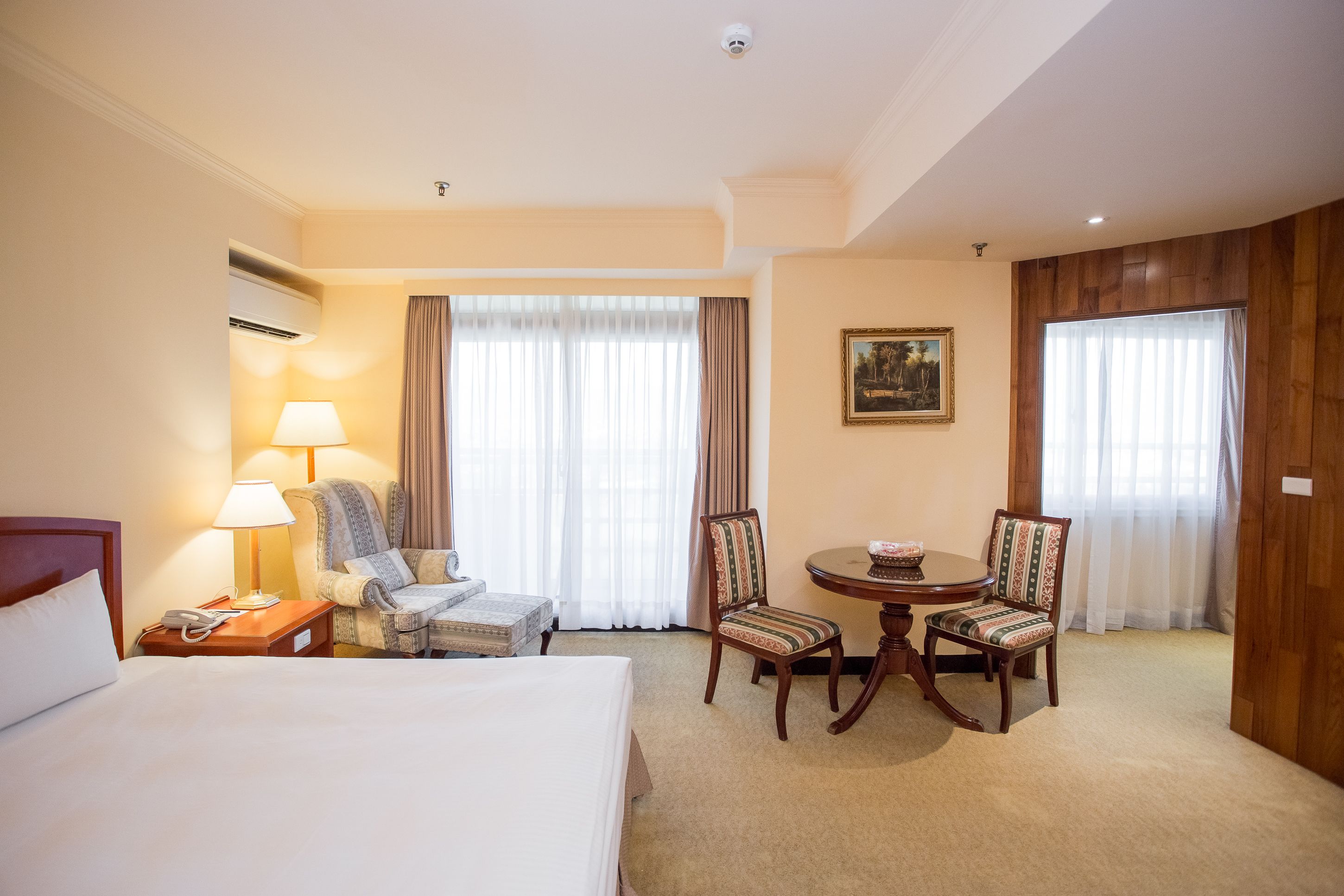 A hotel room with a bed, a desk with a lamp, a chair, a round table with a vase, and two windows with curtains.