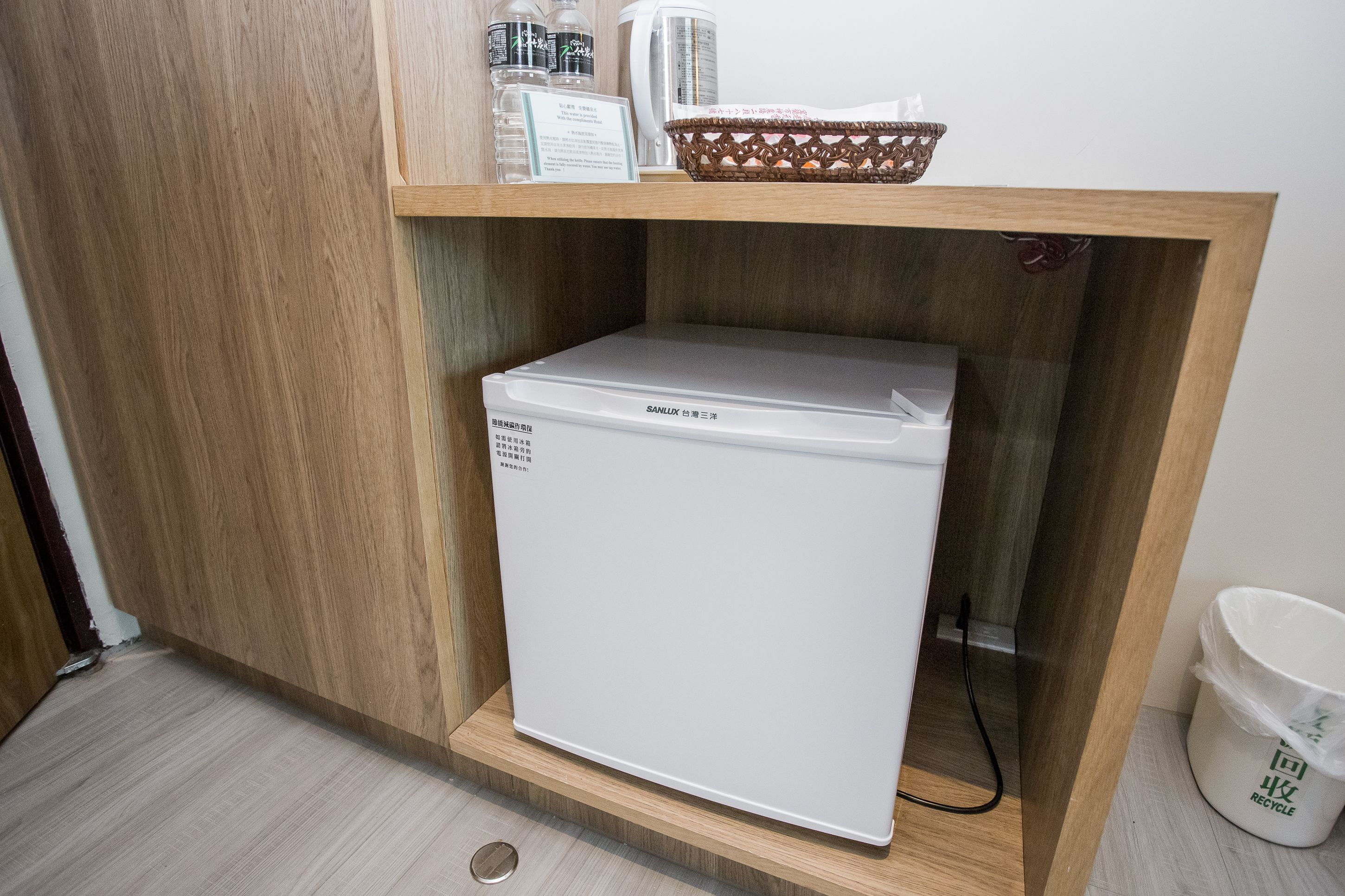 Un petit réfrigérateur blanc, installé dans un meuble en bois, avec un panier et une poubelle de recyclage sur le dessus.