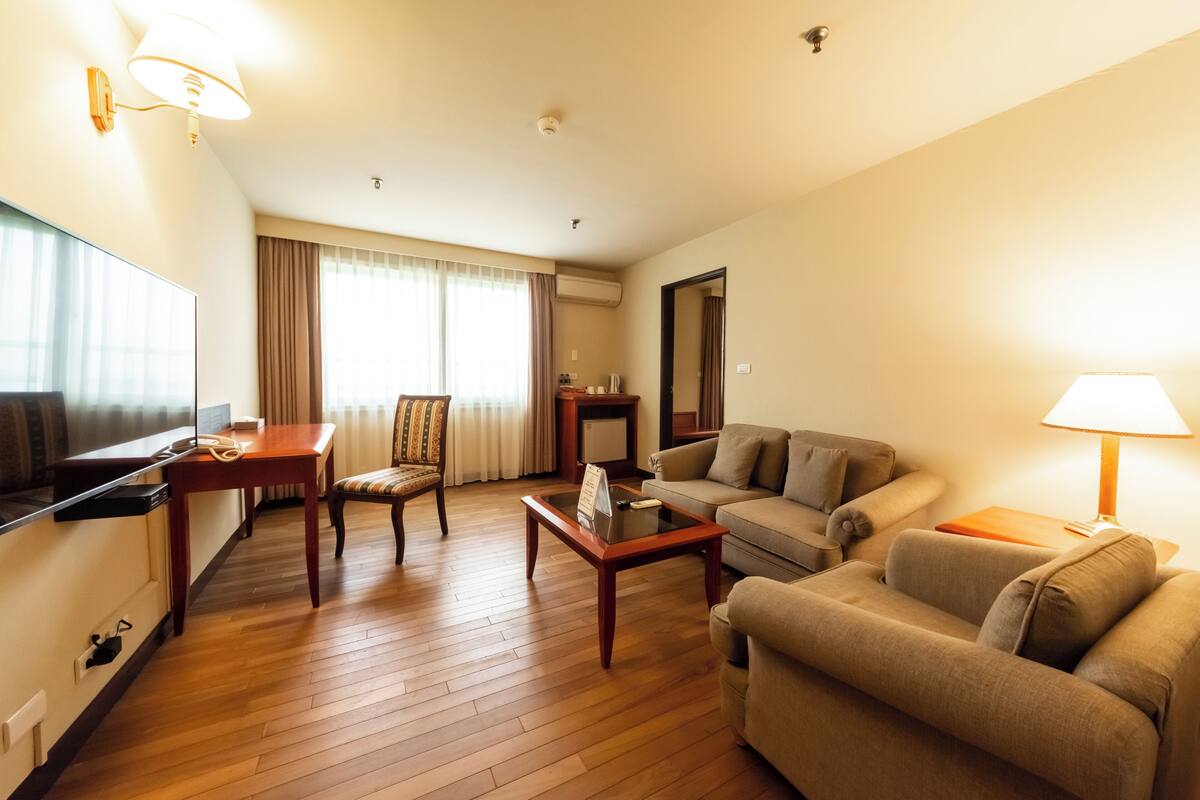 A hotel room with a wooden floor, a sofa, a chair, a coffee table, a TV, a lamp, and a large window with curtains.