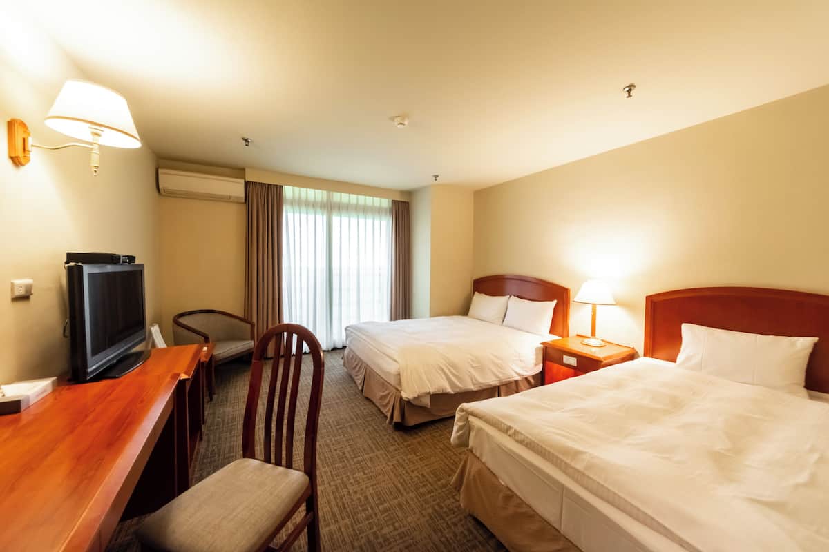A hotel room with two beds, a desk with a television, a chair, a nightstand with a lamp, and a window with curtains.