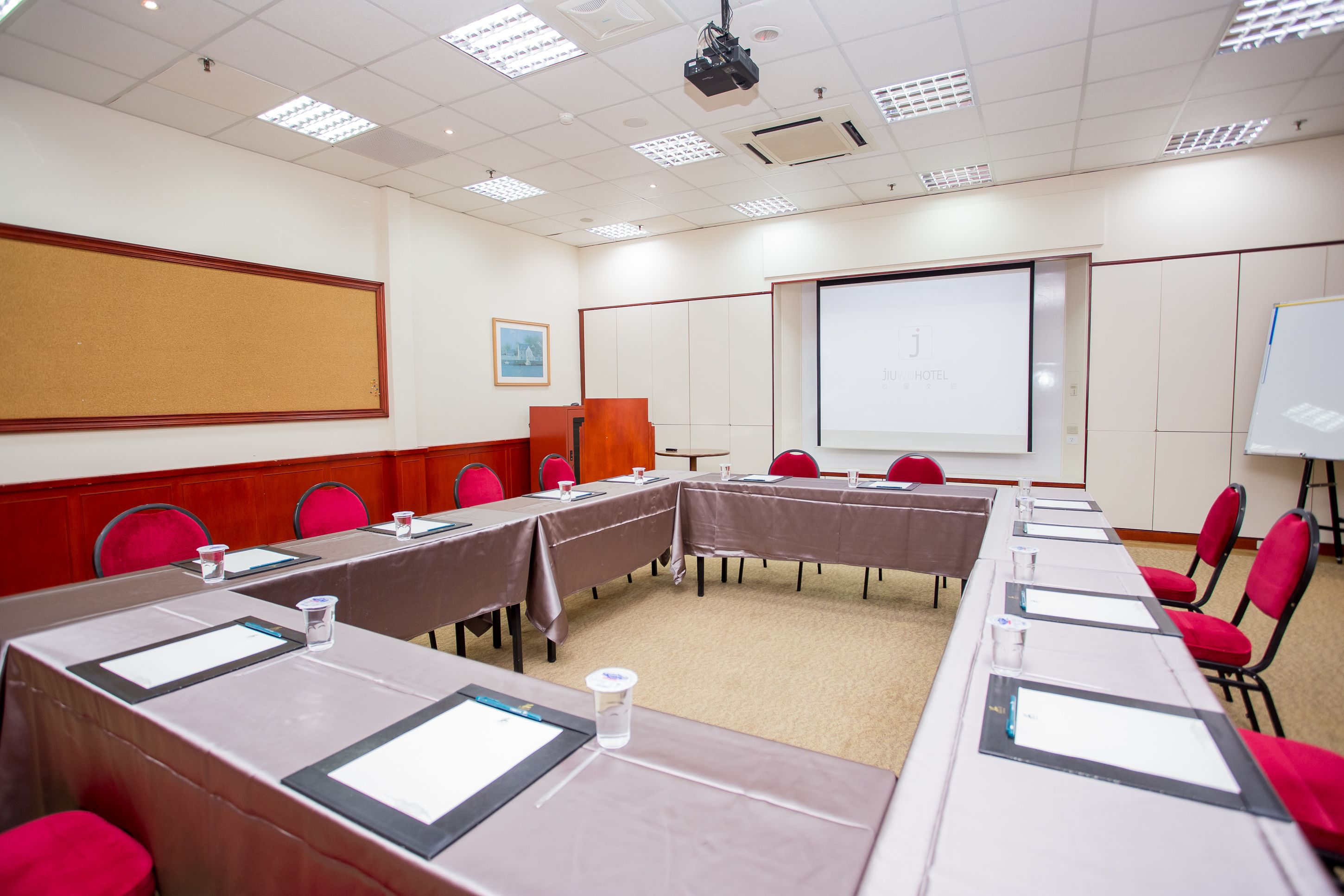 Ein Konferenzraum mit langen Tischen, roten Stühlen, einer Projektionsleinwand und einer Whiteboard-Tafel.