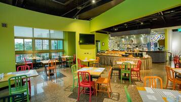 A brightly lit restaurant with colorful chairs and tables, large windows, and a kitchen area visible through an open partition.