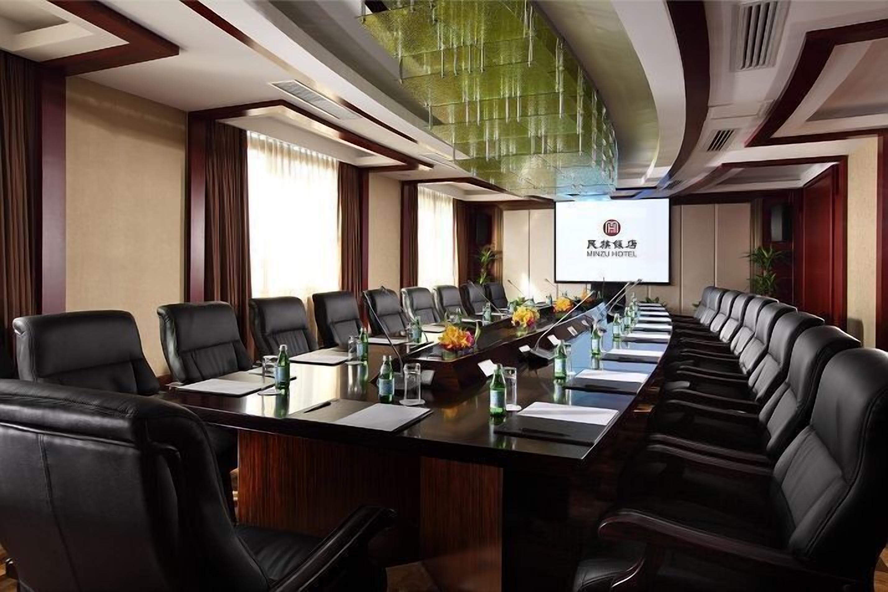 A conference room with a long table, black leather chairs, and a projector screen displaying hotel branding.