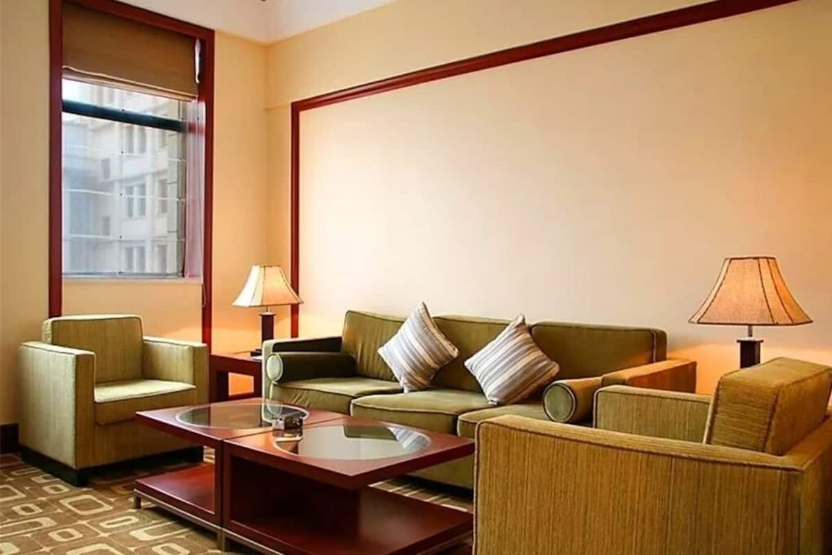 Une chambre d’hôtel avec un canapé vert, une table basse en bois, un fauteuil et une fenêtre avec des stores.