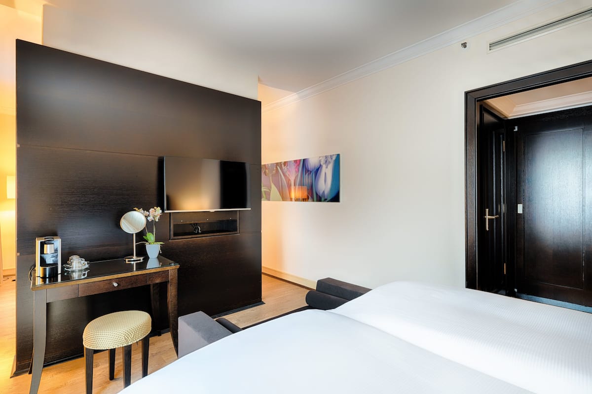 A hotel room with a bed, a desk with a chair, a TV, and a painting on the wall.