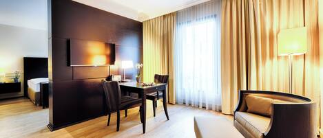 A modern hotel room with a flat-screen TV, a dining table with chairs, a sofa, and a large window with curtains.