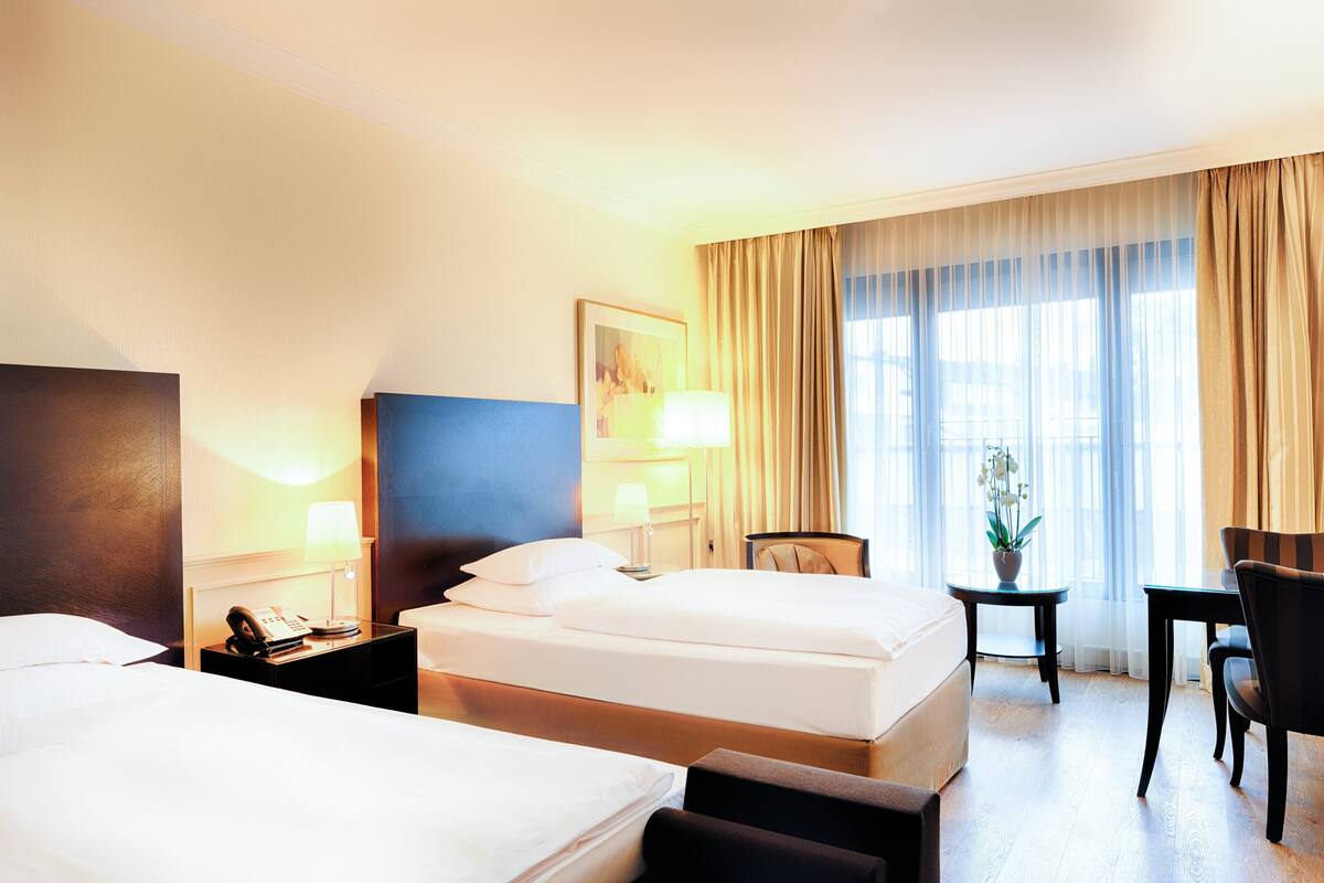 A hotel room with two beds, a desk, a chair, a lamp, and a window with curtains.