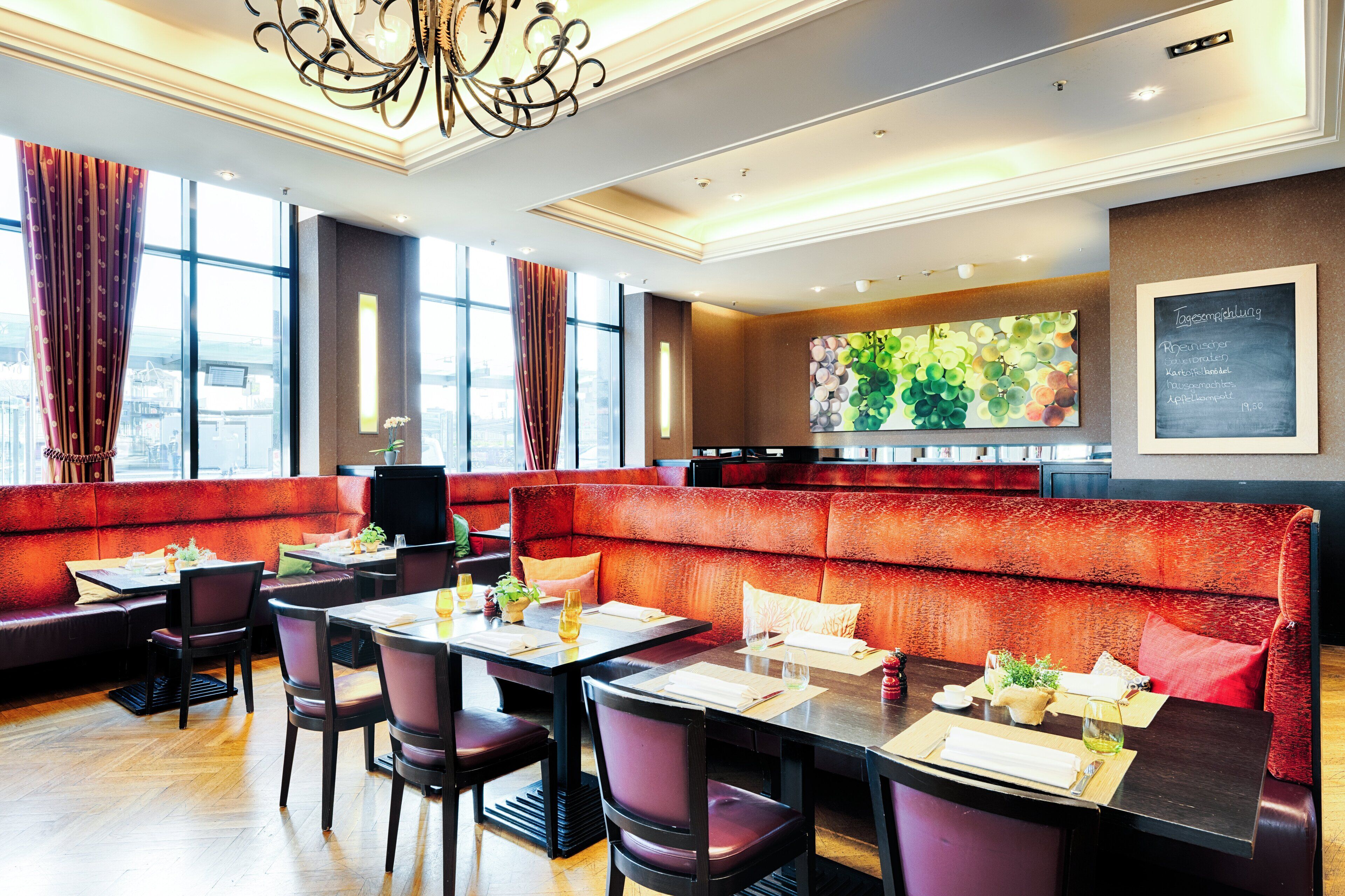 Un restaurante con asientos tapizados en rojo, mesas y sillas de madera. Cuenta con ventanas amplias con cortinas, un candelabro y un panel de menú fijado a la pared.