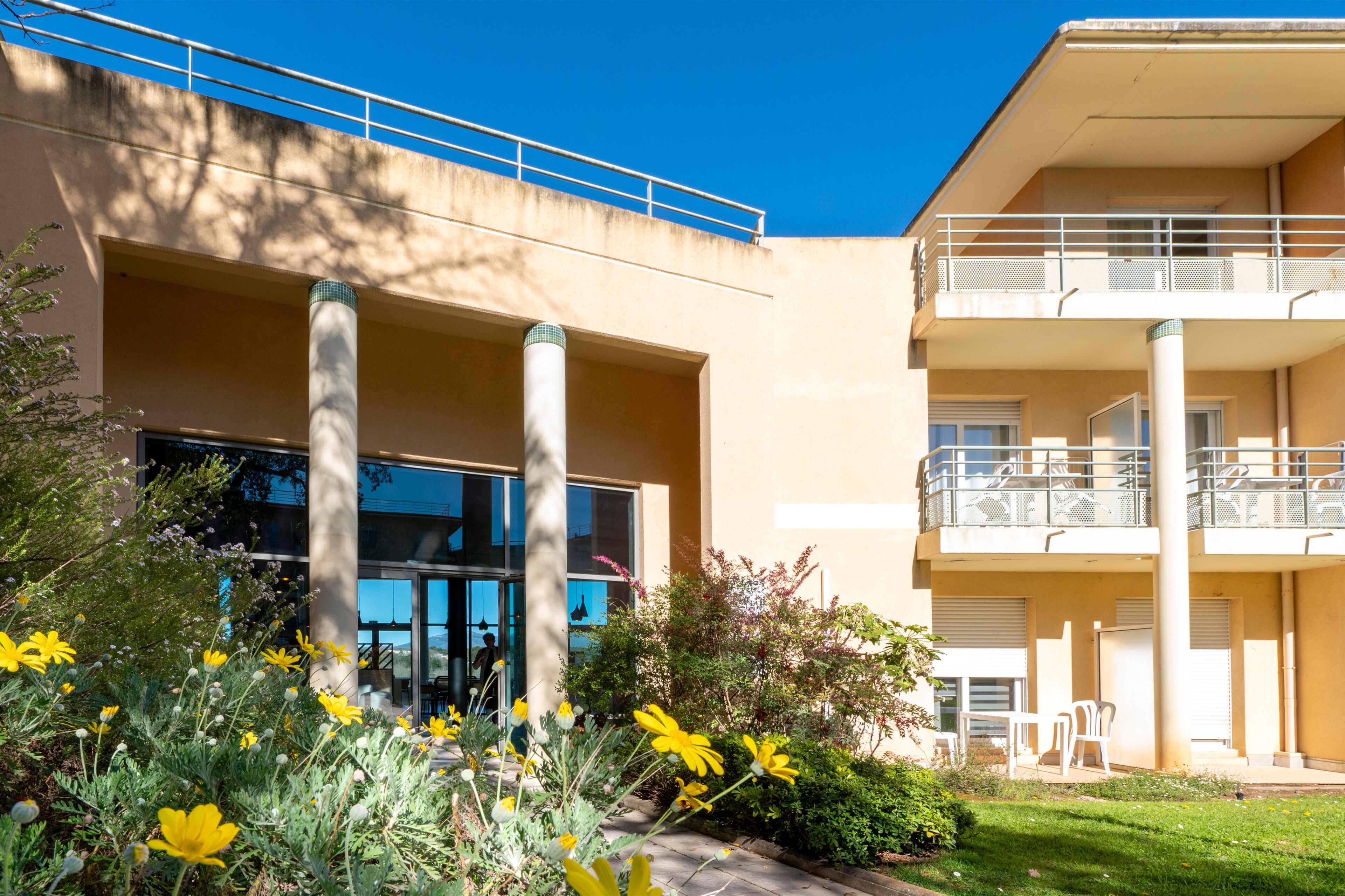 Een modern gebouw met een balkon, een glazen entree en een tuin met gele bloemen.