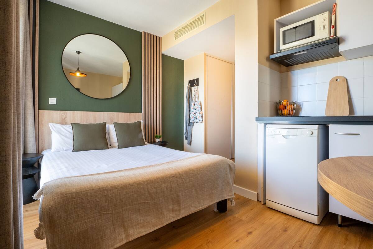 Un dormitorio con cama, mesita de noche, espejo, cómoda y una kitchenette con microondas y refrigerador pequeño.