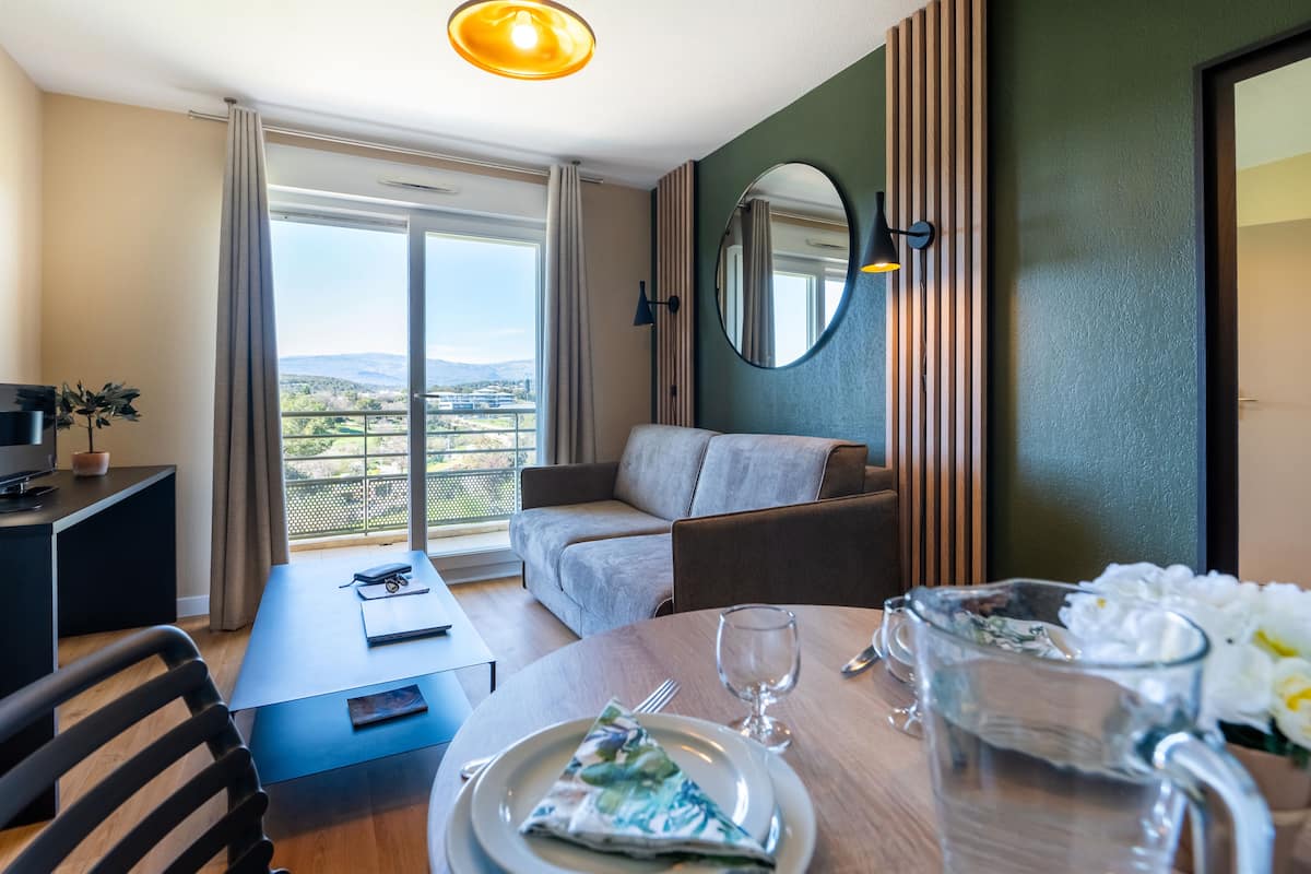 Habitación de hotel con mesa de comedor preparada para dos personas, un sofá, un ventanal con vistas y un espejo en la pared.