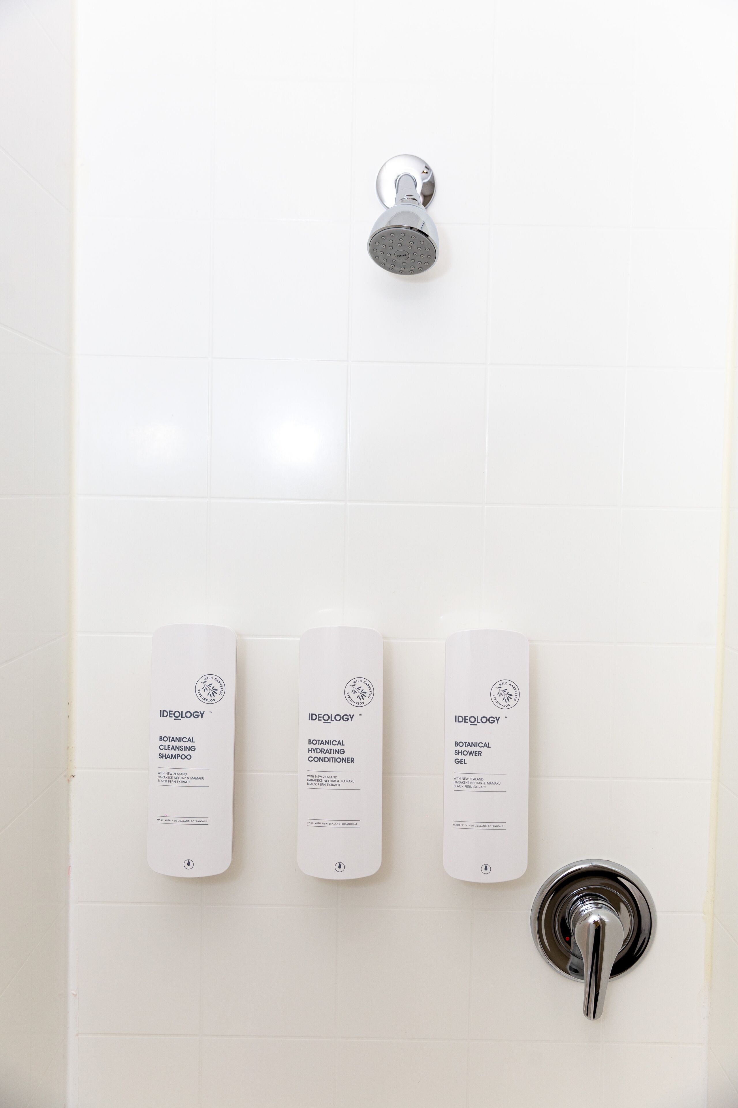 Un baño con ducha, tres botellas de champú, acondicionador y gel de ducha Ideology botanical, y un grifo moderno.